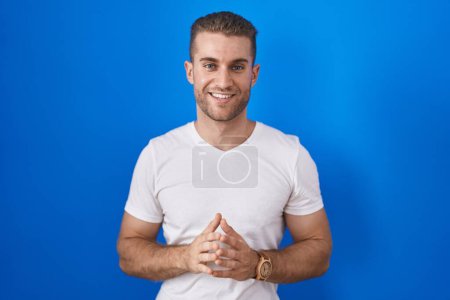 Young caucasian man standing over blue background hands together and fingers crossed smiling relaxed and cheerful. success and optimistic  Poster 640430178