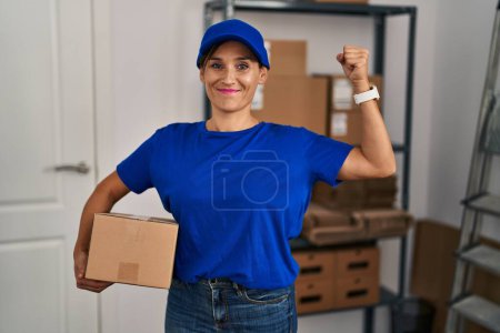 Téléchargez les photos : Femme brune d'âge moyen travaillant en uniforme de livraison et casquette personne forte montrant muscle du bras, confiante et fière de la puissance - en image libre de droit