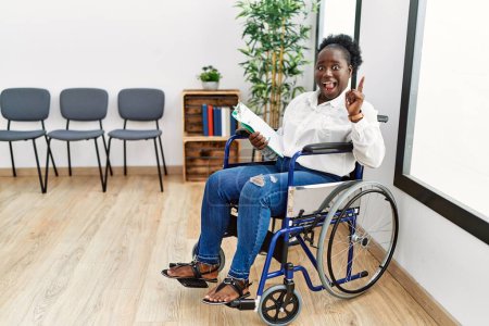 Téléchargez les photos : Jeune femme noire assise en fauteuil roulant dans la salle d'attente pointant du doigt avec une idée réussie. Sorti et heureux. numéro un. - en image libre de droit