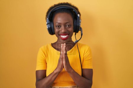 Photo for African woman with curly hair standing over yellow background wearing headphones praying with hands together asking for forgiveness smiling confident. - Royalty Free Image