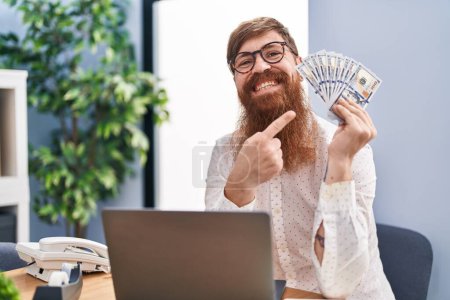 Téléchargez les photos : Homme caucasien avec une longue barbe de travail à l'aide d'un ordinateur portable tenant des dollars souriant heureux pointant avec la main et le doigt - en image libre de droit
