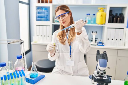 Téléchargez les photos : Jeune femme blonde portant un uniforme scientifique versant du liquide à l'aide d'une pipette au laboratoire - en image libre de droit