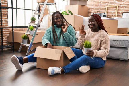 Foto de Dos mujeres africanas se mudan a una nueva casa sentadas en el suelo haciendo buen signo con los dedos, sonriendo gesto amistoso excelente símbolo - Imagen libre de derechos