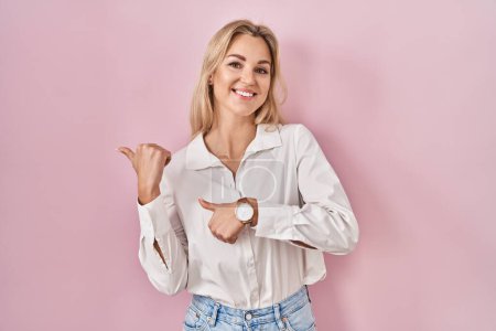 Foto de Mujer caucásica joven con camisa blanca casual sobre fondo rosa apuntando a la espalda con la mano y los pulgares hacia arriba, sonriendo confiado - Imagen libre de derechos