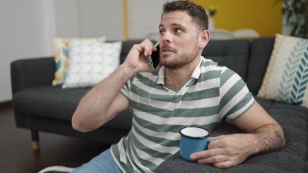 Photo for Young caucasian man speaking on the phone sitting on the sofa at home - Royalty Free Image