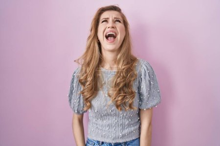 Foto de Beautiful blonde woman standing over pink background angry and mad screaming frustrated and furious, shouting with anger. rage and aggressive concept. - Imagen libre de derechos