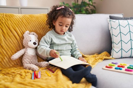 Photo for Adorable hispanic girl drawing on notebook sitting on sofa at home - Royalty Free Image