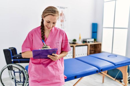 Photo for Young caucasian woman wearing physiotherapist uniform writing on checklist standing at physiotherapy clinic - Royalty Free Image