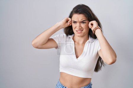Foto de Young teenager girl standing over white background covering ears with fingers with annoyed expression for the noise of loud music. deaf concept. - Imagen libre de derechos
