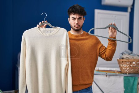 Photo for Hispanic man with beard holding sweater on hanger at laundry room skeptic and nervous, frowning upset because of problem. negative person. - Royalty Free Image