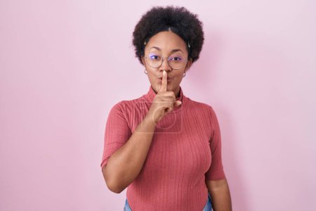Téléchargez les photos : Beautiful african woman with curly hair standing over pink background asking to be quiet with finger on lips. silence and secret concept. - en image libre de droit