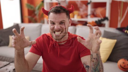 Photo for Young caucasian man screaming wearing devil costume sitting on the sofa at home - Royalty Free Image