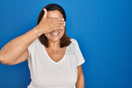 Photo for Hispanic mature woman standing over blue background smiling and laughing with hand on face covering eyes for surprise. blind concept. - Royalty Free Image
