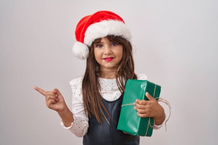 Photo for Little hispanic girl wearing christmas hat and holding gifts smiling happy pointing with hand and finger to the side - Royalty Free Image