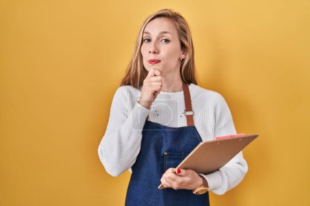 Photo for Young blonde woman wearing professional waitress apron holding clipboard serious face thinking about question with hand on chin, thoughtful about confusing idea - Royalty Free Image