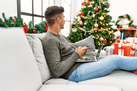 Foto de Joven hombre hispano usando laptop y smartphone sentado en el sofá por el árbol de Navidad en casa - Imagen libre de derechos