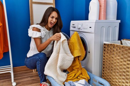 Foto de Mujer hispana joven poniendo ropa sucia en la lavadora enojada y loca gritando frustrada y furiosa, gritando con ira. rabia y concepto agresivo. - Imagen libre de derechos