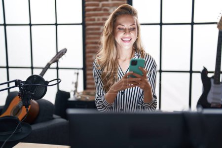 Téléchargez les photos : Jeune femme blonde musicienne souriante confiante en utilisant un smartphone au studio de musique - en image libre de droit