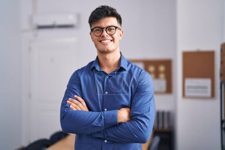 Foto de Joven trabajador de negocios hispano sonriendo confiado de pie con los brazos cruzados gesto en la oficina - Imagen libre de derechos