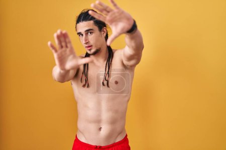 Téléchargez les photos : Homme hispanique avec les cheveux longs debout torse nu sur fond jaune faisant cadre à l'aide de mains paumes et doigts, perspective de la caméra - en image libre de droit