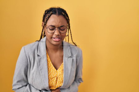 Téléchargez les photos : Femme afro-américaine avec des tresses debout sur fond jaune avec la main sur le ventre parce que l'indigestion, la maladie douloureuse se sent mal. concept de douleur. - en image libre de droit