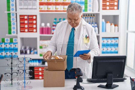 Téléchargez les photos : Homme d'âge moyen aux cheveux gris pharmacien utilisant le pavé tactile tenant bouteille de pilules à la pharmacie - en image libre de droit