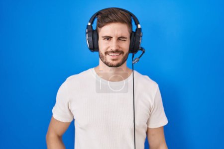 Téléchargez les photos : Homme hispanique avec barbe écoutant de la musique portant des écouteurs cligner des yeux en regardant la caméra avec une expression sexy, gai et heureux visage. - en image libre de droit
