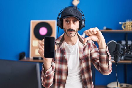 Photo for Hispanic man with beard at music studio showing smartphone with angry face, negative sign showing dislike with thumbs down, rejection concept - Royalty Free Image