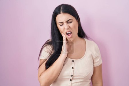 Foto de Young hispanic woman standing over pink background touching mouth with hand with painful expression because of toothache or dental illness on teeth. dentist - Imagen libre de derechos