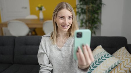 Photo for Young blonde woman taking selfie picture with smartphone sitting on the sofa at home - Royalty Free Image