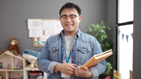 Photo for Young chinese man teacher smiling confident holding books at kindergarten - Royalty Free Image