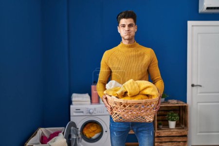 Foto de Young hispanic man holding laundry basket puffing cheeks with funny face. mouth inflated with air, catching air. - Imagen libre de derechos