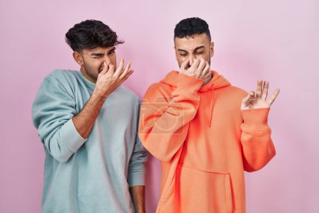 Foto de Young hispanic gay couple standing over pink background smelling something stinky and disgusting, intolerable smell, holding breath with fingers on nose. bad smell - Imagen libre de derechos
