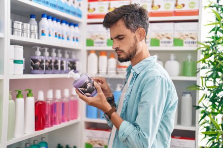 Téléchargez les photos : Young hispanic man customer reading label gel hands bottle at pharmacy - en image libre de droit