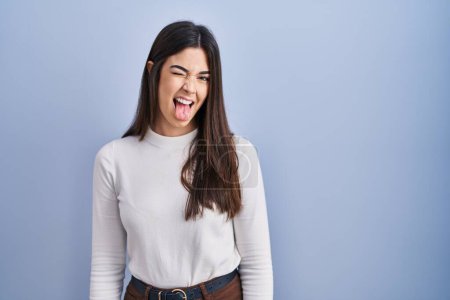 Photo for Young brunette woman standing over blue background sticking tongue out happy with funny expression. emotion concept. - Royalty Free Image