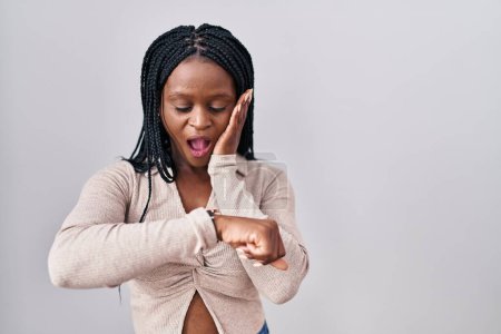 Téléchargez les photos : Femme africaine avec des tresses debout sur fond blanc regardant le temps de la montre inquiet, peur de se faire tard - en image libre de droit