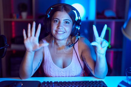 Foto de Mujer rubia joven jugando videojuegos con auriculares que muestran y señalan hacia arriba con los dedos número ocho mientras sonríe confiado y feliz. - Imagen libre de derechos