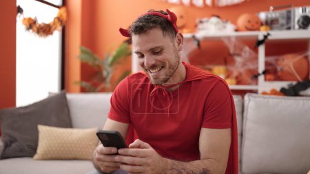 Téléchargez les photos : Jeune homme caucasien portant costume diable assis sur le canapé en utilisant smartphone à la maison - en image libre de droit