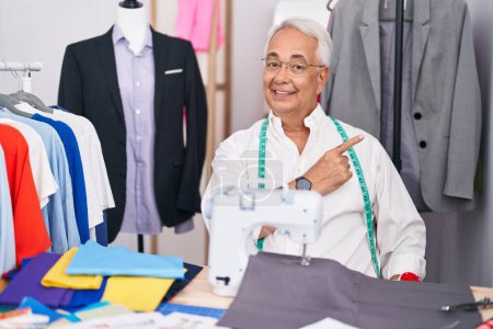 Photo for Middle age man with grey hair dressmaker using sewing machine with a big smile on face, pointing with hand finger to the side looking at the camera. - Royalty Free Image