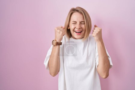 Téléchargez les photos : Jeune femme caucasienne debout sur fond rose excité pour le succès avec les bras levés et les yeux fermés célébrant la victoire en souriant. concept gagnant. - en image libre de droit
