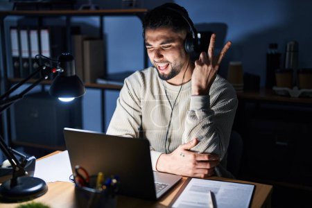 Photo for Young handsome man working using computer laptop at night smiling with happy face winking at the camera doing victory sign. number two. - Royalty Free Image