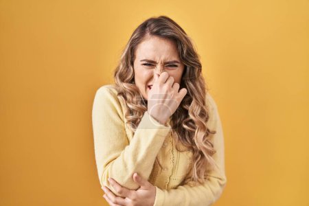 Foto de Young caucasian woman standing over yellow background smelling something stinky and disgusting, intolerable smell, holding breath with fingers on nose. bad smell - Imagen libre de derechos