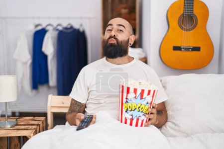 Junger hispanischer Mann mit Bart und Tätowierungen, der Popcorn im Bett isst und in die Kamera schaut, die einen Kuss bläst, der schön und sexy ist. Liebesausdruck. 