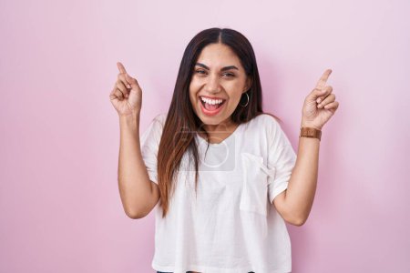 Photo for Young arab woman standing over pink background smiling amazed and surprised and pointing up with fingers and raised arms. - Royalty Free Image