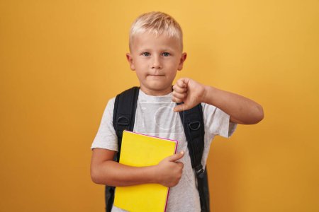 Photo for Little caucasian boy wearing student backpack and holding book with angry face, negative sign showing dislike with thumbs down, rejection concept - Royalty Free Image