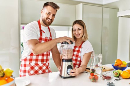 Téléchargez les photos : Jeune couple souriant confiant faire smoothie en utilisant mélangeur à la cuisine - en image libre de droit