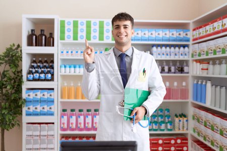 Téléchargez les photos : Jeune homme hispanique travaillant à la pharmacie pharmacie tenant stéthoscope souriant heureux pointant avec la main et le doigt vers le côté - en image libre de droit