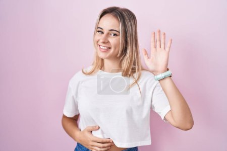 Téléchargez les photos : Young blonde woman standing over pink background waiving saying hello happy and smiling, friendly welcome gesture - en image libre de droit