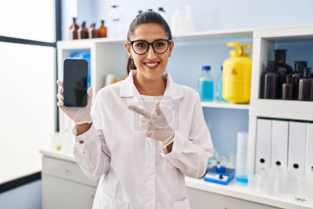 Téléchargez les photos : Jeune femme hispanique travaillant au laboratoire scientifique avec smartphone souriant heureux pointant avec la main et le doigt - en image libre de droit