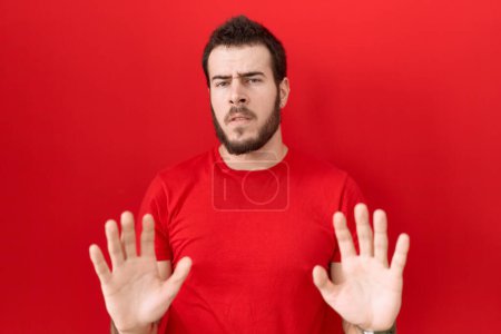 Foto de Joven hombre hispano con camiseta roja casual alejando las palmas de las manos mostrando rechazo y negación con expresión de miedo y repugnante. parada y prohibido. - Imagen libre de derechos